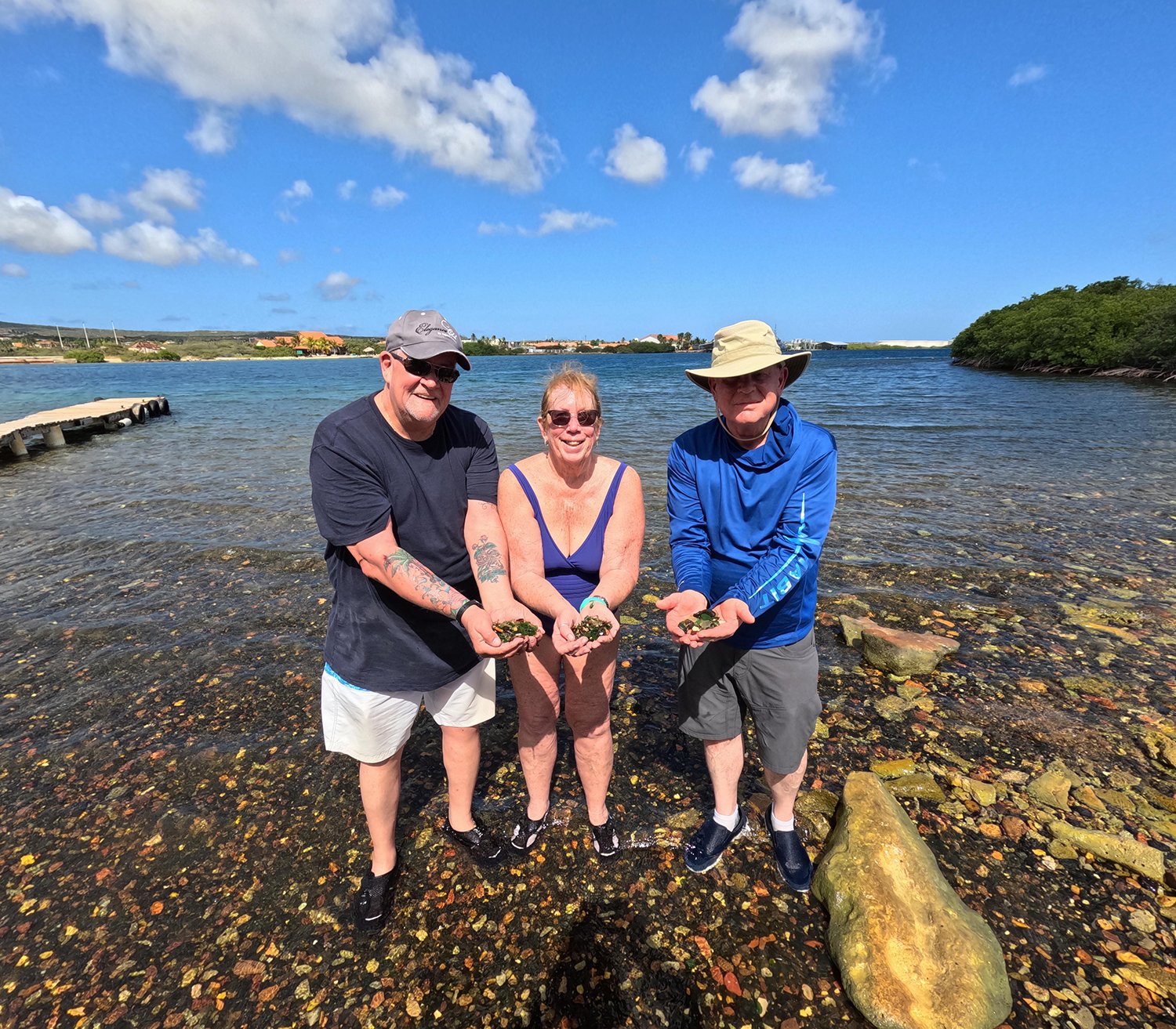 Three smiling tourists display their sea glass finds on a shallow shore in Aruba.